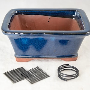 May include: A blue ceramic bonsai pot with a brown clay interior. The pot has a rectangular shape with rounded corners and a slightly flared lip. The pot is sitting on a white surface with two black wire mesh squares and a black wire coil.