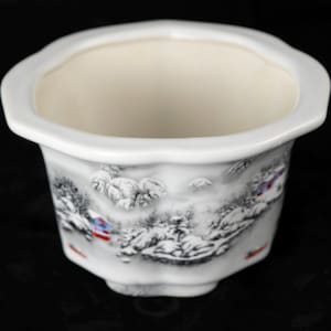 May include: White ceramic planter with a scalloped rim and a winter landscape design. The planter features a detailed black and white illustration of a snowy mountain scene with small houses and trees. The interior is a cream color.