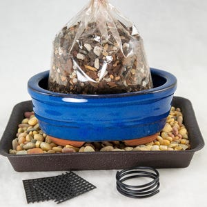 May include: A blue ceramic bonsai pot with a bag of soil inside, sitting on a brown plastic tray with small stones. A black plastic grid and a black wire coil are also visible.