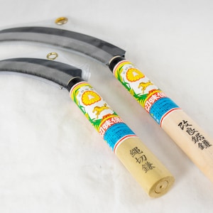 May include: Two Japanese gardening tools with wooden handles. The handles are wrapped in paper with Japanese characters and a yellow flower design. The blades are curved and have a serrated edge.
