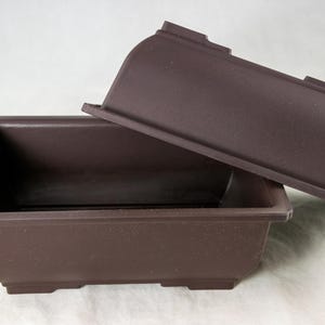 May include: A rectangular dark brown plastic bonsai pot with a wide rim and a matching dark brown plastic tray positioned at an angle resting against the back edge of the pot. The container features a smooth matte finish and four small feet.