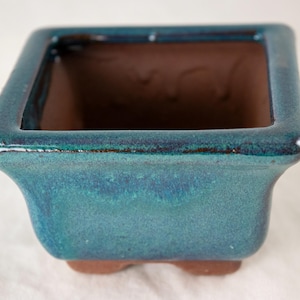 May include: A square, teal glazed ceramic bonsai pot with a brown interior. The pot has a slightly flared rim and a rounded base.