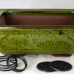 May include: A green glazed ceramic bonsai pot with a rectangular shape and a brown interior. The pot has a speckled green glaze and is sitting on a white surface. There are two black plastic drainage mats and three black metal rings in the image.