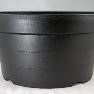 May include: A black plastic plant pot with a textured surface. The pot has a wide, flat rim and a series of horizontal bands around the body. The base has small feet. Ideal for indoor or outdoor plants.