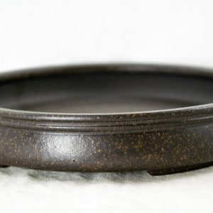 May include: A brown ceramic bonsai pot with a rounded shape and a slightly flared rim. The pot has a textured surface and three small feet on the bottom.