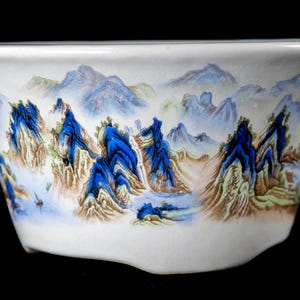May include: White ceramic bowl with a painted landscape scene. The bowl features a detailed depiction of blue mountains, trees, and water, with brown and green accents. The design is set against a white background.
