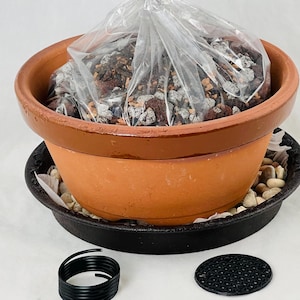 May include: A terracotta pot with a black saucer, filled with a clear plastic bag of potting soil. The pot is sitting on a white surface. There is a black wire coil and a black plastic mesh disc on the surface.