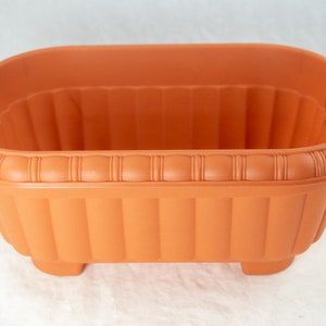 May include: A terracotta-colored rectangular planter with a decorative rim and ribbed sides. The planter has four small feet.