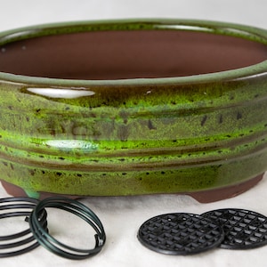 May include: A green glazed oval bonsai pot with a brown rim and a textured surface. The pot is sitting on a white surface with two black plastic drainage mats and three metal rings.