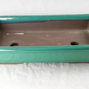 May include: A rectangular green glazed bonsai pot with two drainage holes. The pot has a brown interior and a light green rim.