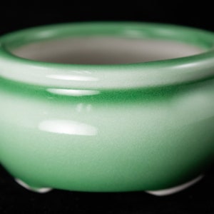 May include: A green ceramic pot with a white interior. The pot has a rounded shape and a smooth, glossy finish.