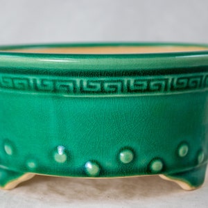 May include: A green ceramic planter with a decorative border and small raised dots around the bottom. The planter has three feet.