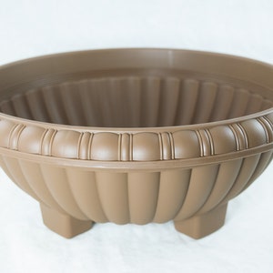 May include: A brown plastic planter with a fluted design and a decorative rim. The planter has three legs and is suitable for indoor or outdoor use.