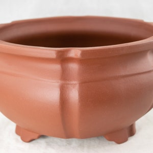 May include: A terracotta-colored ceramic pot with a rounded base and four feet. The pot has a wide, rounded opening and a smooth, slightly textured surface.