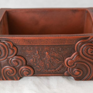 May include: A rectangular terracotta bonsai pot with a brown glaze. The pot has a raised design of clouds and figures on the front. The pot is empty.