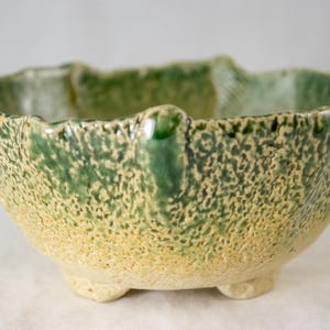 May include: A ceramic bowl with a textured, uneven rim and a footed base. The exterior features a mottled glaze with a gradient of earthy yellow at the bottom transitioning into a vibrant green toward the top, creating a rustic, organic appearance.