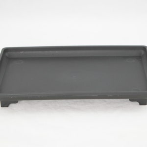 May include: A dark gray plastic plant tray with a rectangular shape and raised edges. The tray has four small feet on the bottom.
