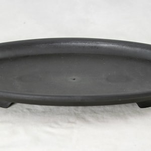 May include: Black oval-shaped plant saucer with three small feet. The saucer is made of plastic and has a drainage hole in the center.