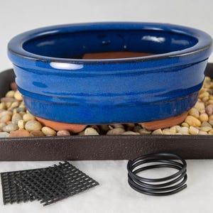 May include: A blue oval bonsai pot with a brown tray, a black plastic grid, and a black wire coil. The pot is sitting on top of the tray, which is filled with small stones.