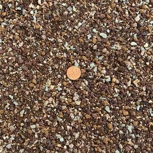 May include: Close-up of a brown, red, and white soil mix with a penny for scale. The soil is a blend of small rocks, pebbles, and dirt.