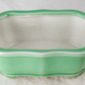 May include: A light green ceramic bonsai pot with a white rim and a slightly rounded rectangular shape.