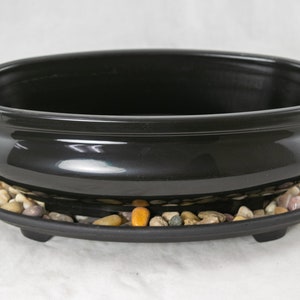 May include: A black oval ceramic bonsai pot with a matching black plastic saucer. The saucer has small decorative stones in it.