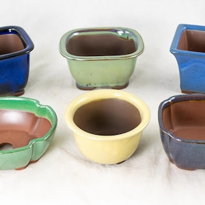 May include: Six ceramic bonsai pots in various colors and shapes. The pots are arranged in two rows of three. The pots are all glazed and have a smooth finish.