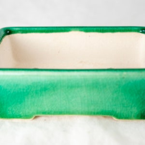 May include: A rectangular green ceramic bonsai pot with a white interior. The pot has a slightly raised rim and a smooth, glazed finish.