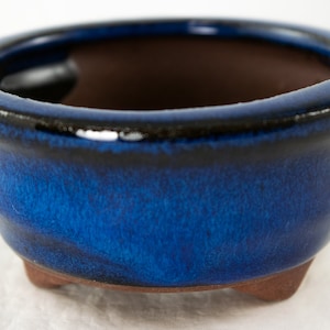 May include: A small, blue ceramic bonsai pot with a black rim and three small feet. The pot is glazed with a deep blue color and has a slight sheen.