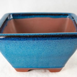 May include: A square bonsai pot with a glossy blue exterior and a brown interior. The pot has a dark blue rim and sits on four small, brown feet. The ceramic pot is designed for planting small trees or plants.