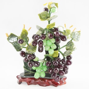 May include: A jade grapevine sculpture with green leaves and purple grapes on a wooden base. The sculpture is made of jade and is a traditional Chinese art form.