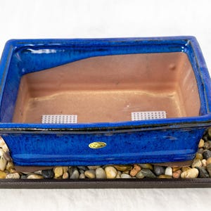 May include: A blue ceramic bonsai pot with a brown interior sits on a black plastic tray filled with small stones. The pot has two small white mesh screens inside.
