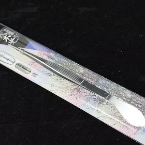May include: A silver stainless steel tweezers with a flat, rounded tip. The tweezers are packaged in a clear plastic sleeve with a colorful background and the text "YAGIMITSU" printed on the bottom right corner.