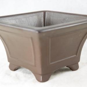 May include: A square brown ceramic planter with four legs. The planter has a smooth, matte finish.