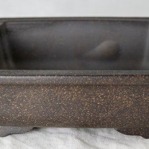 May include: A square, dark brown ceramic bonsai pot with a rough texture and four small feet.