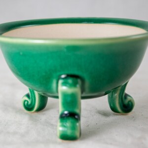 May include: A green ceramic bowl with three legs. The bowl has a white interior and a glossy finish.