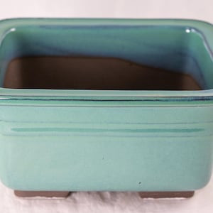 May include: A square, teal-colored ceramic bonsai pot with a brown interior and four small feet.