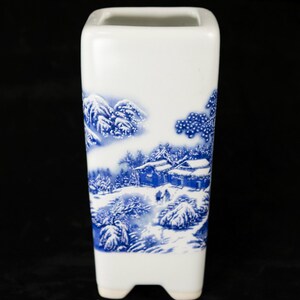 May include: A white ceramic vase with a rectangular shape and a blue and white winter landscape design. The scene depicts snow-covered mountains, trees, and buildings. The vase has a square opening and three small feet.