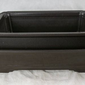 May include: Two black plastic rectangular bonsai pots, one nested inside the other. The larger pot is slightly wider than the smaller pot.