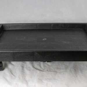 May include: A black, rectangular rolling plant stand. The stand has a textured surface and sits on four black caster wheels. The stand is designed to hold a plant pot or other items. The stand is made of a durable material.