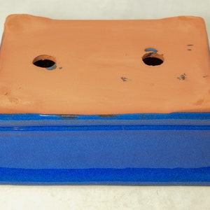 May include: A blue glazed rectangular ceramic planter with two drainage holes in the bottom. The planter is made of terracotta and has a blue glaze on the outside.