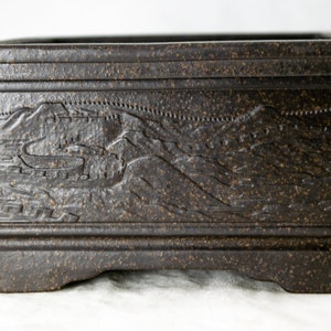 May include: A dark brown square ceramic bonsai pot with a raised design of a mountain range and a wall. The pot has a textured surface and a slightly raised base.
