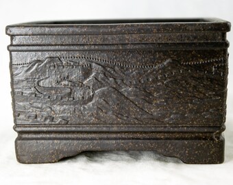 May include: A dark brown square ceramic bonsai pot with a raised design of a mountain range and a wall. The pot has a textured surface and a slightly raised base.