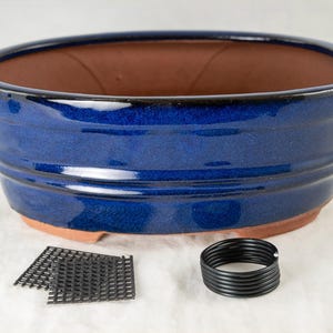 May include: A blue ceramic bonsai pot with a brown clay interior. The pot has a rounded oval shape and a slightly flared rim. It is sitting on a white surface with two black plastic mesh squares and a black plastic coil.