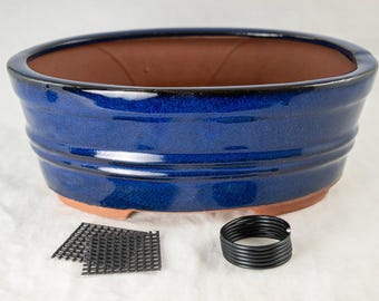 May include: A blue ceramic bonsai pot with a brown clay interior. The pot has a rounded oval shape and a slightly flared rim. It is sitting on a white surface with two black plastic mesh squares and a black plastic coil.