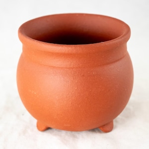May include: A small terracotta pot with three legs. The pot is a reddish-brown color and has a smooth, slightly textured surface.