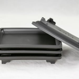 May include: Four black plastic square trays with raised edges and four small feet on the bottom. The trays are stacked on top of each other.
