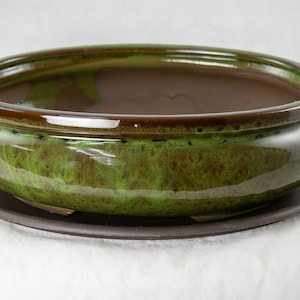 May include: A green glazed ceramic bonsai pot with a brown rim and a matching brown saucer. The pot has a rounded shape and a smooth, glossy finish.