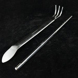 May include: Two stainless steel gardening tools. One tool is a small rake with four tines. The other tool is a long, thin, flat-edged tool with a rounded end.