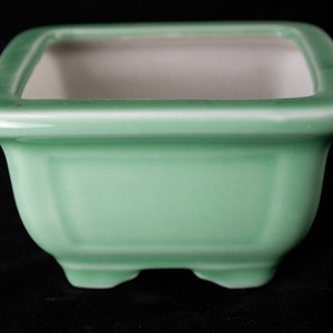 May include: A square, light green ceramic bonsai pot with a white interior and four small feet.